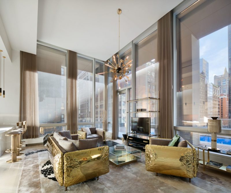 maximalism- contemporary living room, high windows with brown curtains, a brown carpet, cream and gold sofa and armchairs, glass coffee table, a TV, a gold hanging lamp 